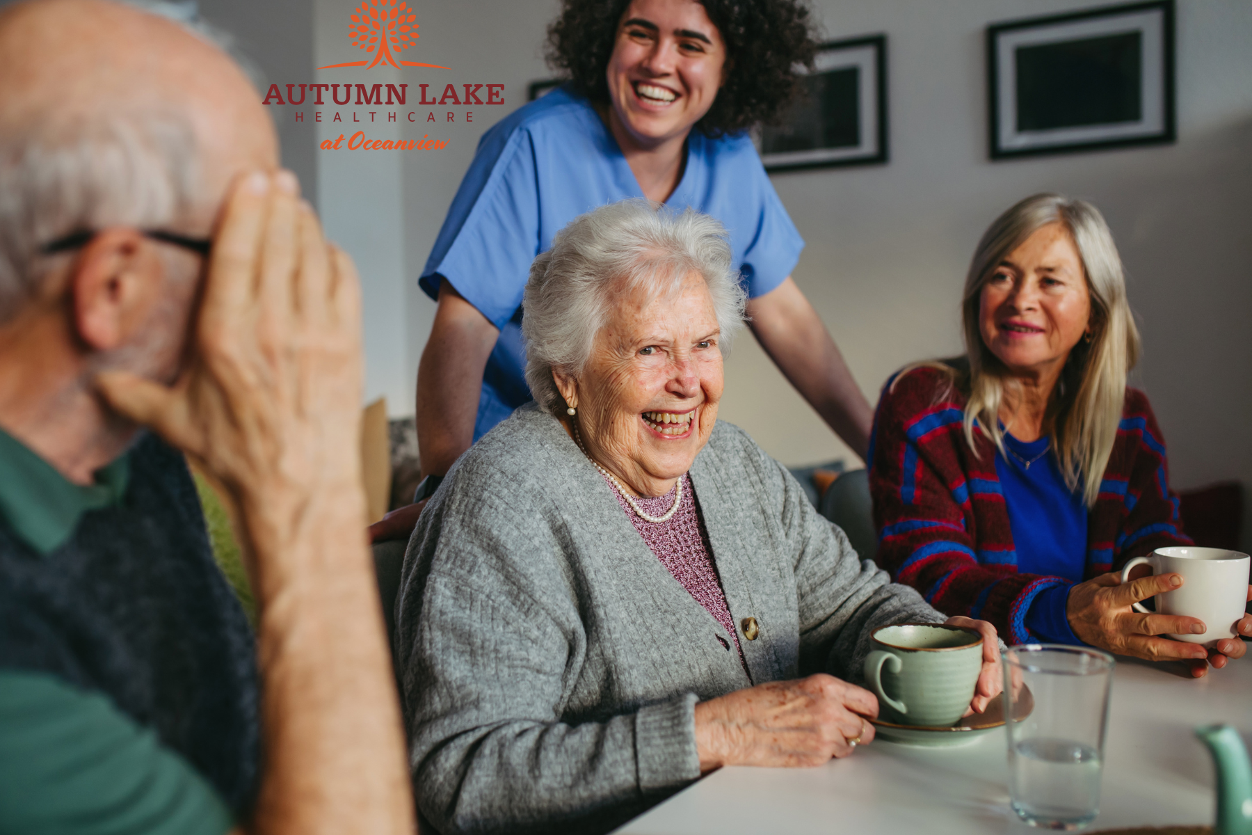 A smiling nurse interacts with a group of seniors enjoying tea and conversation in a nursing home.