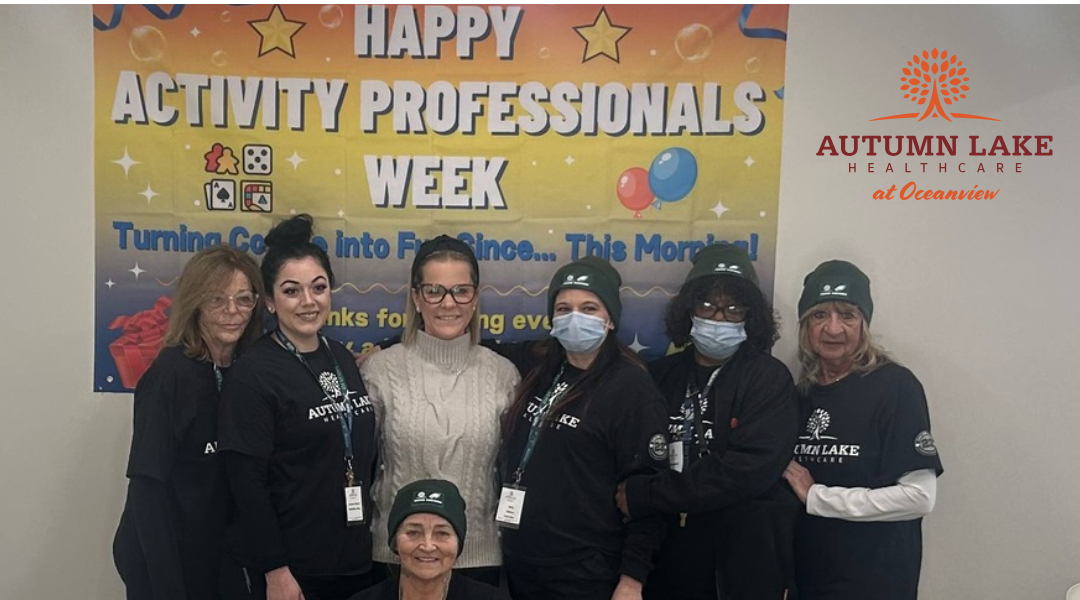 A diverse group of smiling nursing home staff members posing in front of a "Happy Activity Professionals Week" banner.