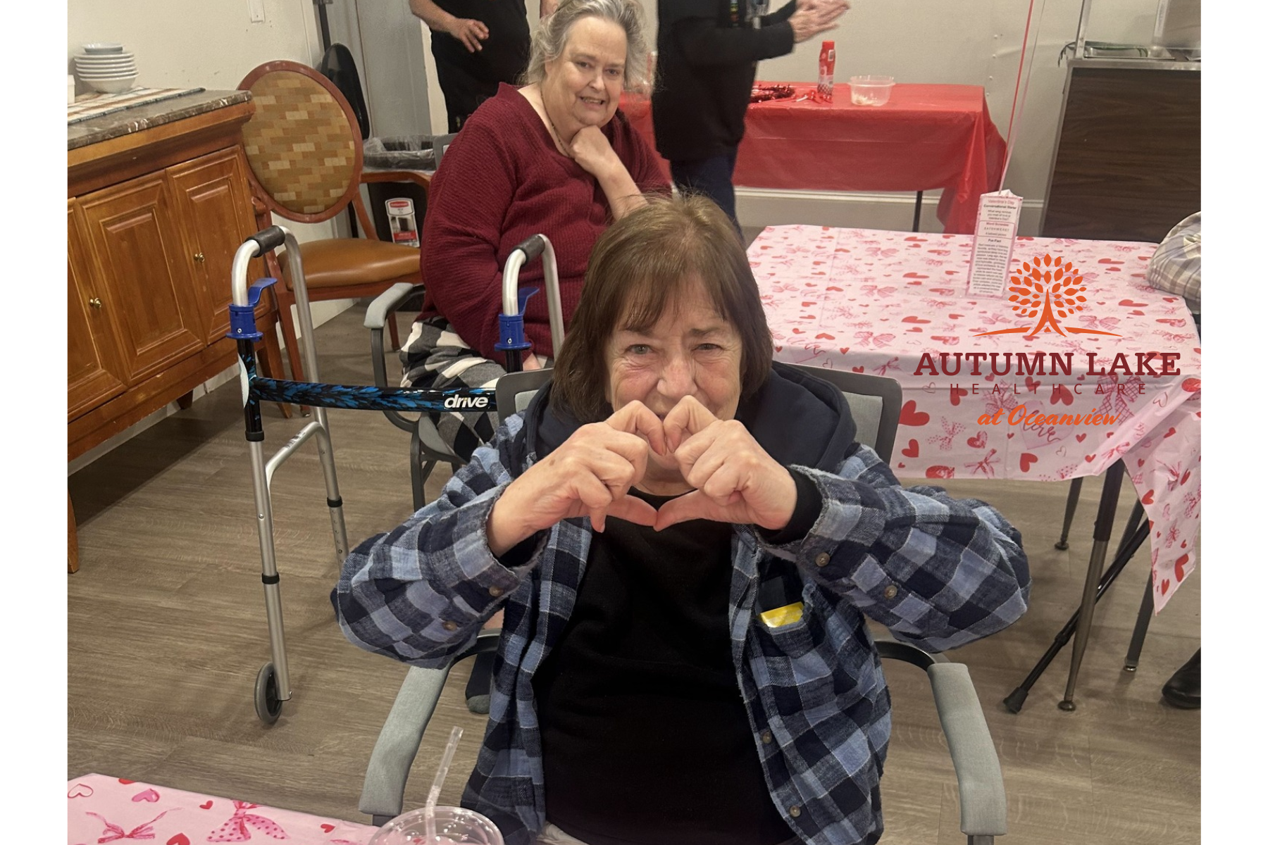 A woman at a rehabilitation center makes a heart shape with her hands while seated at a table decorated for Valentine's Day.