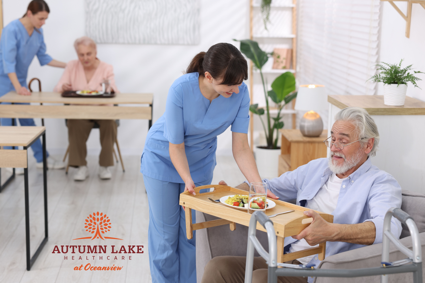 A caregiver in blue scrubs serves a meal to an elderly man in a bright nursing home dining area.