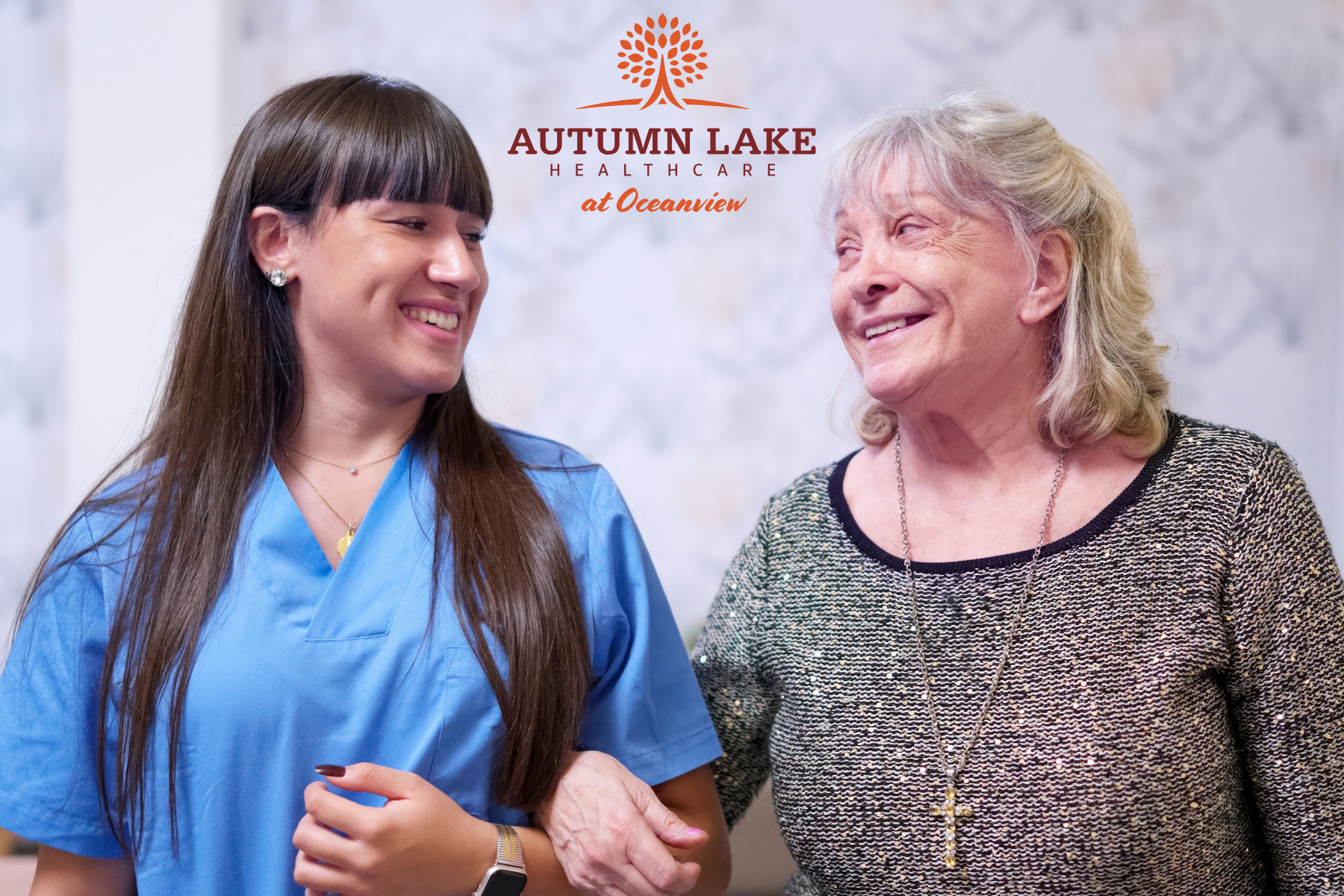 A smiling young nurse in blue scrubs walks arm-in-arm with an elderly woman, both sharing a warm, joyful conversation at Autumn Lake Healthcare at Oceanview.